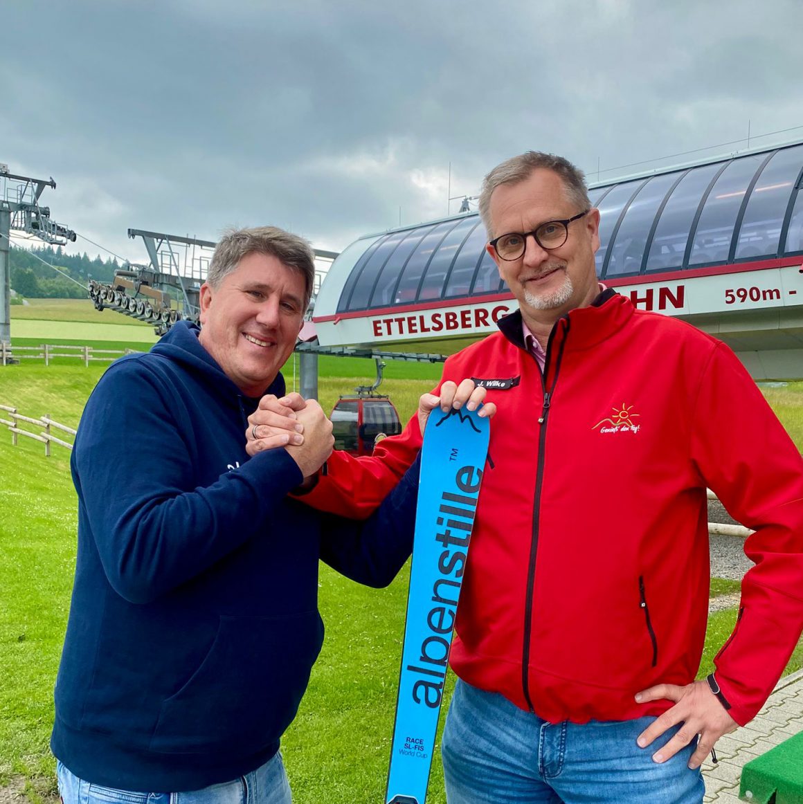 Zwei Männer, einer hält davon einen Skier, stehen vor einer Seilbahnstation