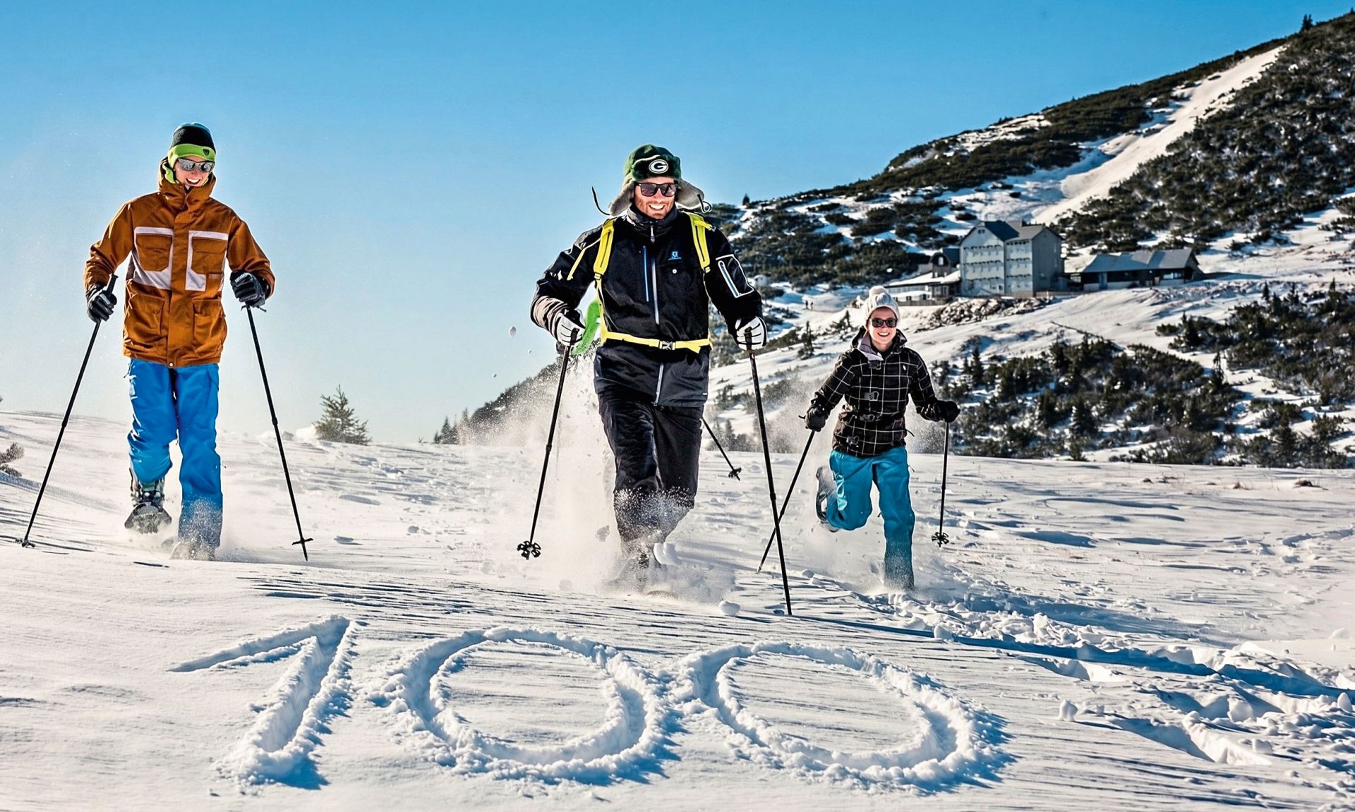 Drei Schneeschuhwanderer laufend im Hintergrund, im Vordergrund ist die Zahl 100 in den Schnee gemalt.